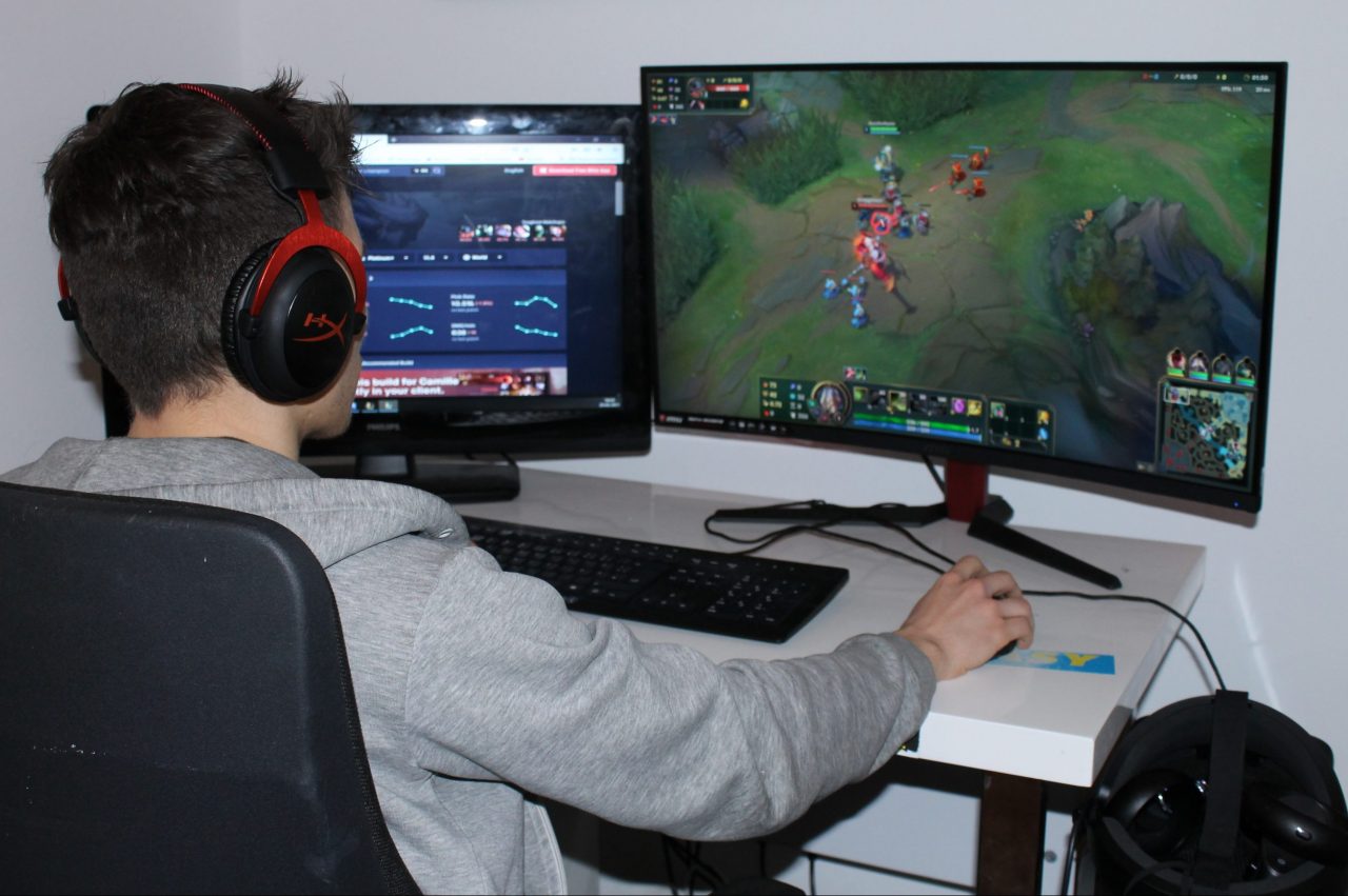 Auf dem Foto ist ein Mann zu sehen, der an einem Schreibtisch sitzt. Vor sich hat er eine Tastatur sowie zwei Computerbildschirme, auf denen ein Spiel zu sehen ist.