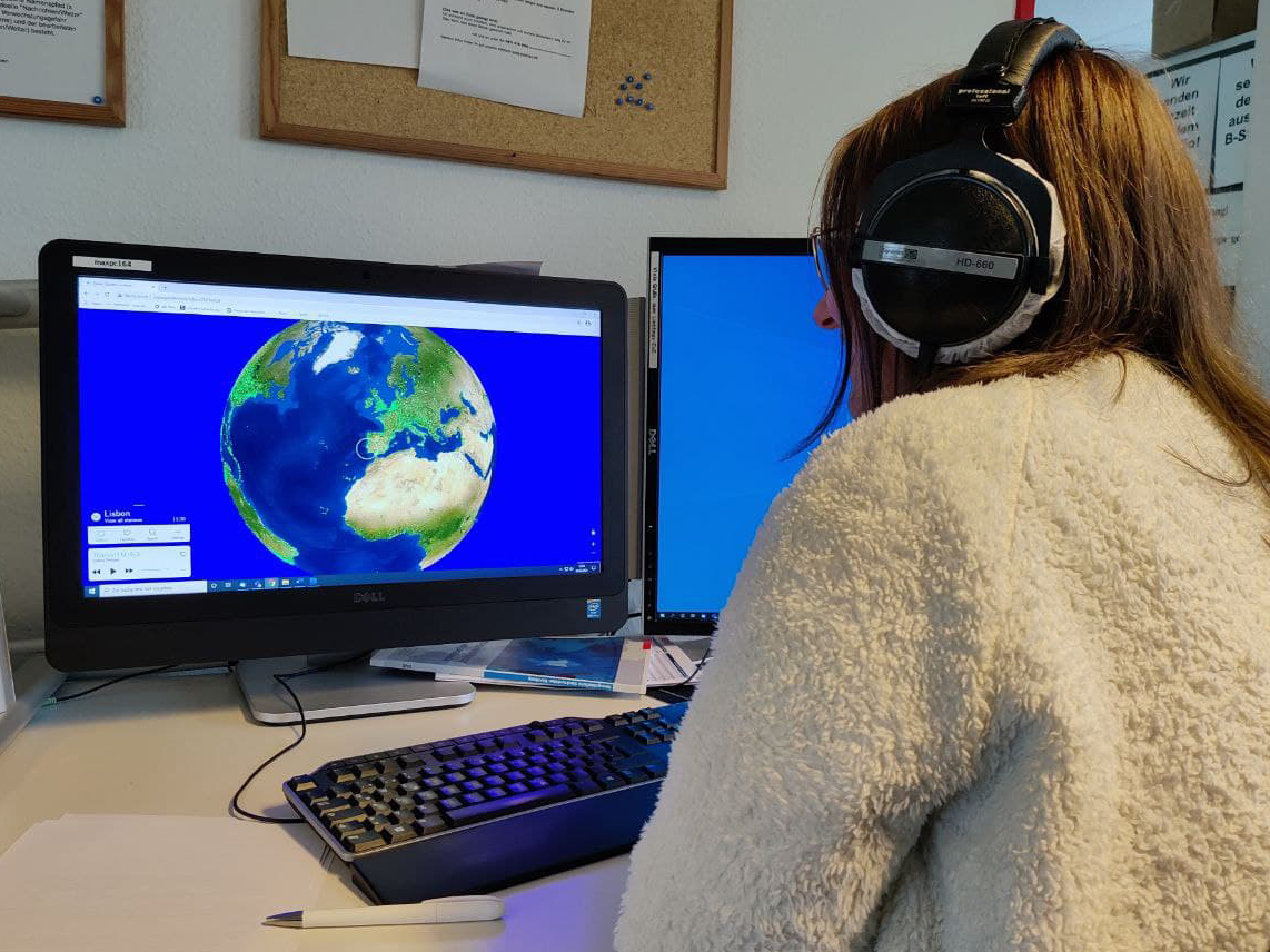 Auf dem Foto ist eine Frau zu sehen, die auf einen Computerbildschirm schaut. Dort ist eine Weltkugel zu sehen, auf der verschiedene Radiostationen eingezeichnet sind.