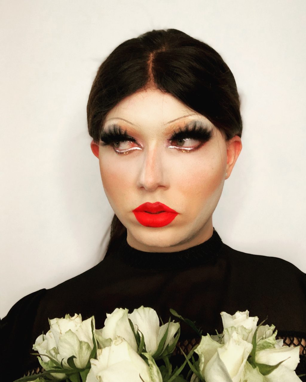 Auf dem Foto ist die Dragqueen Lillet Lube zu sehen. Sie trägt eine schwarze Perücke, lange Wimpern und einen roten Lippenstift. Lillet Lube hält einen Strauß weißer Rosen in der Hand.