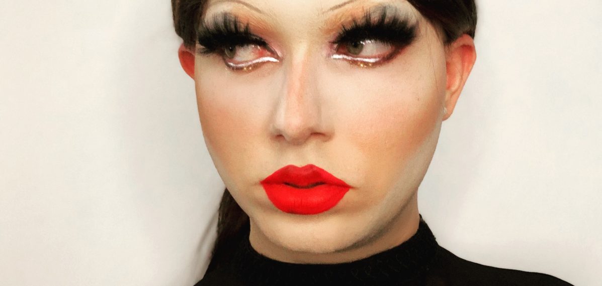 Auf dem Foto ist die Dragqueen Lillet Lube zu sehen. Sie trägt eine schwarze Perücke, lange Wimpern und einen roten Lippenstift. Lillet Lube hält einen Strauß weißer Rosen in der Hand.