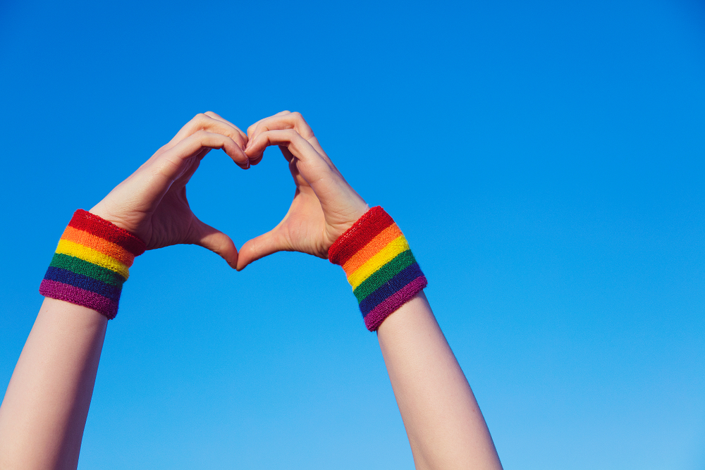 Man sieht zwei Hände, die ein Herz vor einem blauen Himmel bilden. An den Handgelenken befinden sich Schweißbänder in Regenbogenfarben.