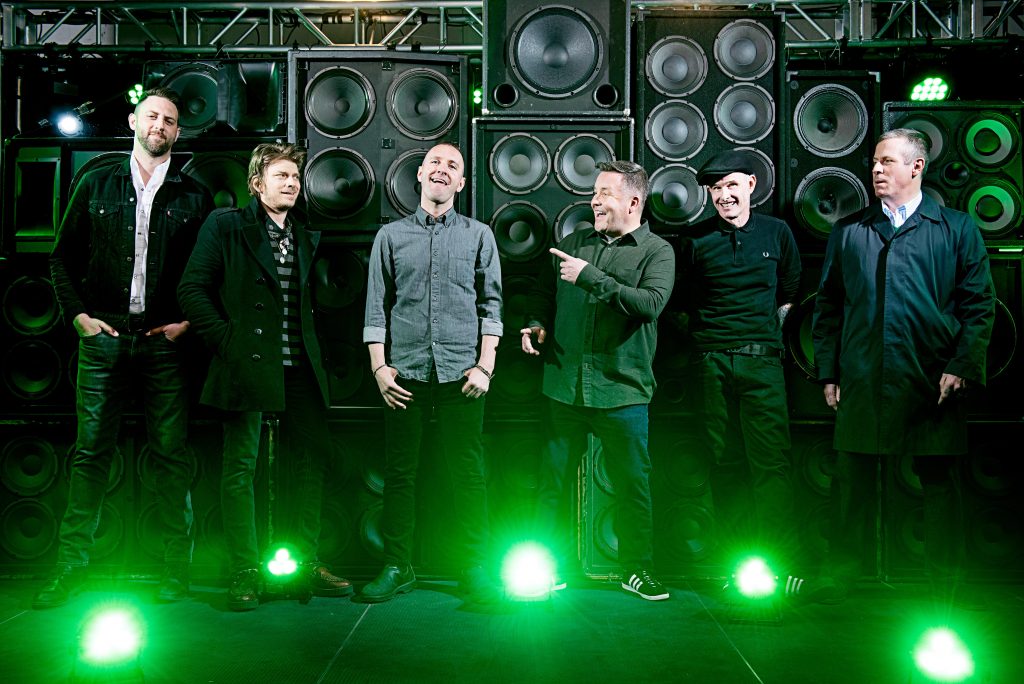 Auf dem Foto sind die Dropkick Murphys zu sehen. Sie stehen vor einer Wand mit Lautsprechern und grünen Scheinwerfern.