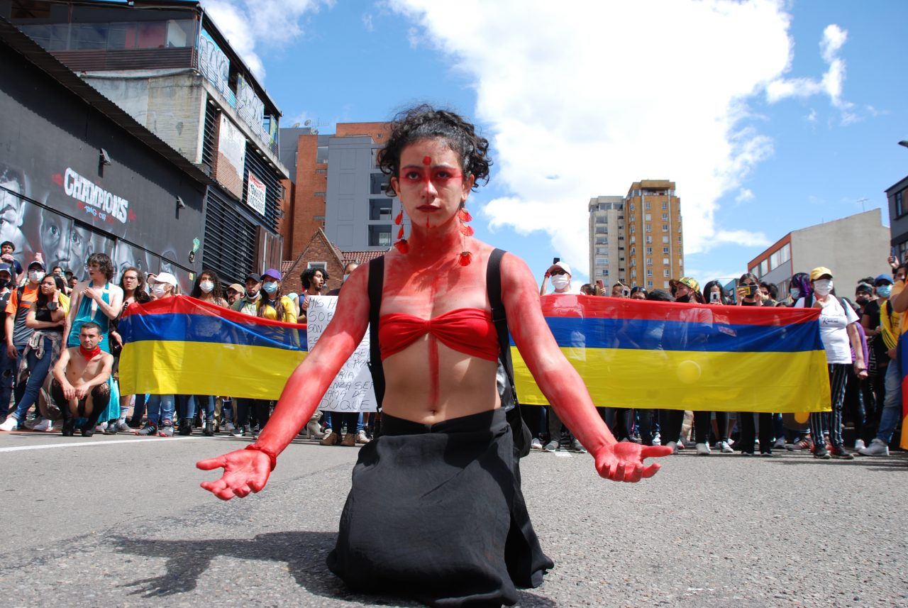 Eine Frau ist rot bemalt und kniet auf der Straße, im Hintergrund stehen Menschen in einer Reihe und halten eine kolumbianische Flagge in der Hand.