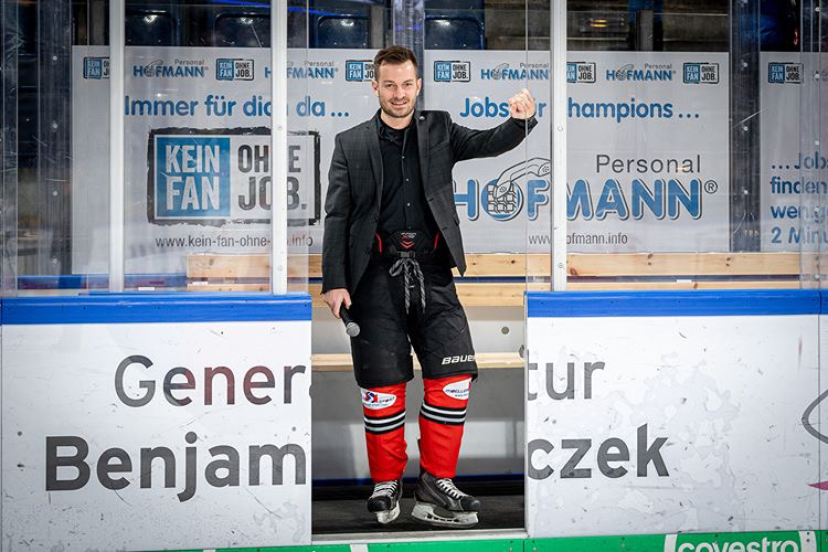 Christian Rupp steht an der Bande der Arena, obenrum mit Hemd und Sakko, untenrum in Eishockey-Montur.