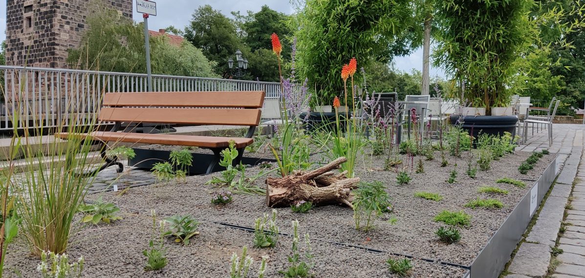 Im Vordergrund sind ein mobiles Blumenbeet und eine Sitzbank zu sehen. Im Hintergrund befinden sich Stühle und Tische von einem Cafe sowie eine Brücke.