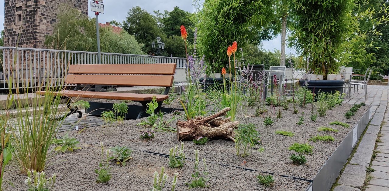 Im Vordergrund sind ein mobiles Blumenbeet und eine Sitzbank zu sehen. Im Hintergrund befinden sich Stühle und Tische von einem Cafe sowie eine Brücke.