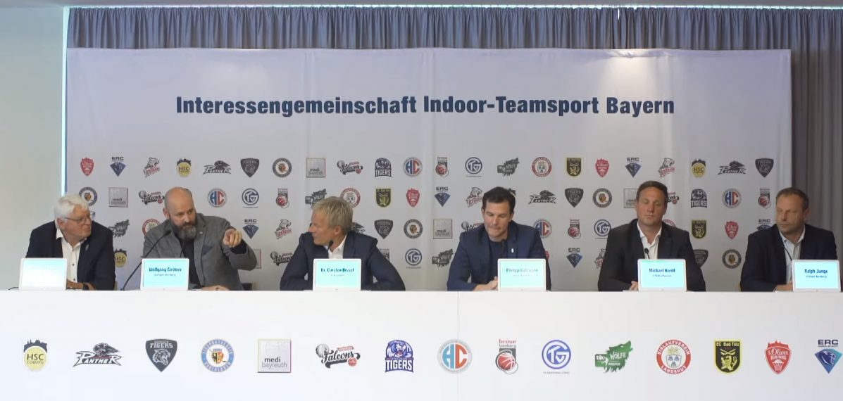 Auf dem Foto sieht man sechs Vertreter der Interessengemeinschaft Indoor-Teamsport Bayern bei einer Pressekonferenz.