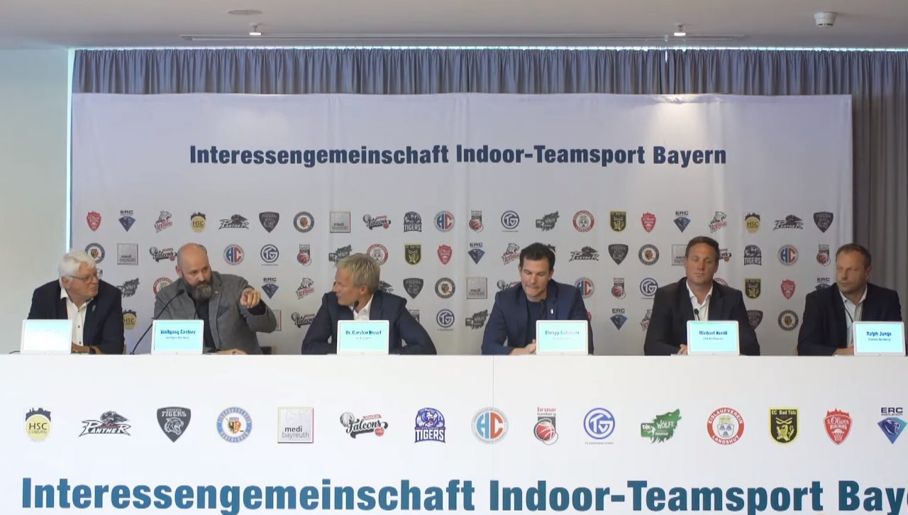 Auf dem Foto sieht man sechs Vertreter der Interessengemeinschaft Indoor-Teamsport Bayern bei einer Pressekonferenz.