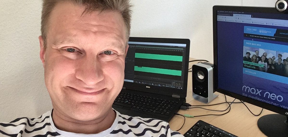 Holger Müller sitzt an seinem Schreibtisch im Home Office. Im Hintergrund des Selfies sind ein PC mit Tastatur und ein Laptop zu sehen.