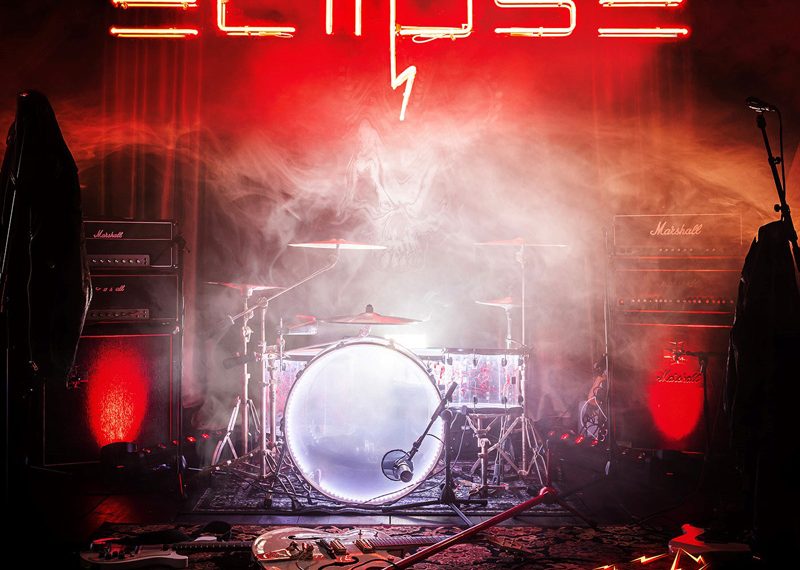 Das Albumcover "Wired" von Eclipse zeigt einen dunklen Raum, in dem ein Schlagzeug, eine Gitarre, ein Mikrofonständer und vieles mehr stehen. Darüber wabern Nebelschwaden und es hängt eine Leuchtschrift mit dem Wort "Eclipse".