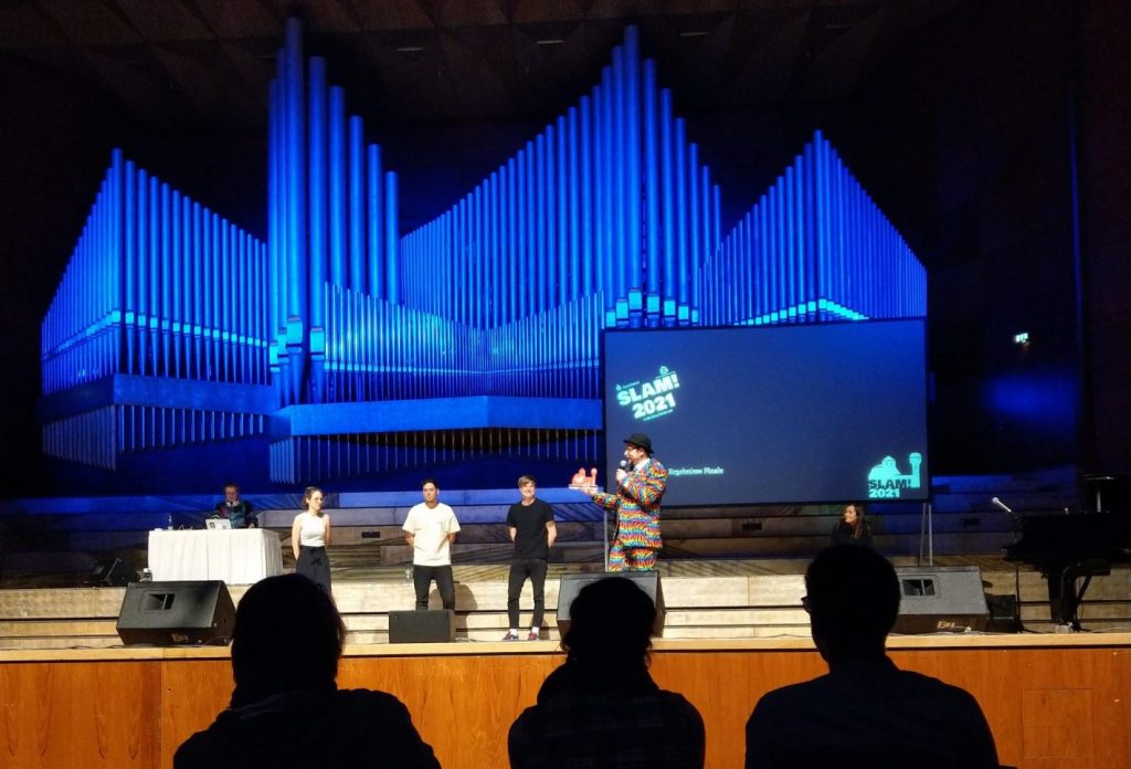 Man sieht die Bühne der Meistersingerhalle, auf der die drei Finalisten und der Moderator mit der Trophäe stehen. Im Hintergrund ist die blau beleuchtete Orgel zu sehen.