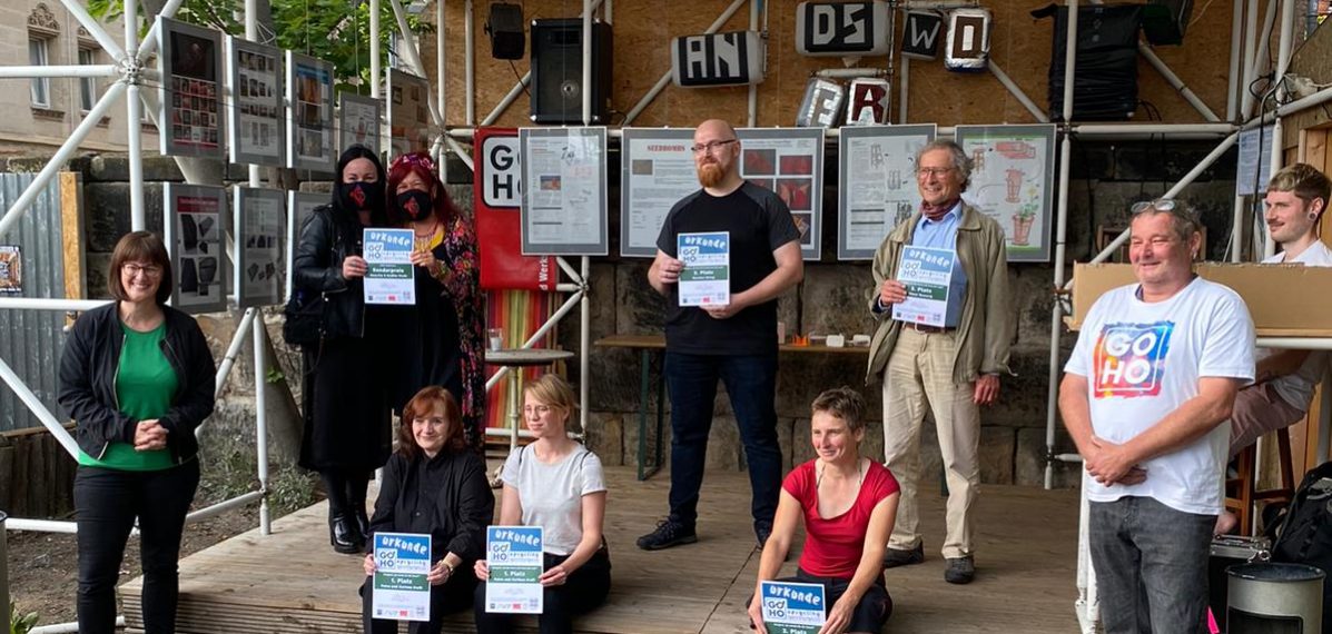 Das Foto zeigt eine Preisverleihung. Es ist ein Gruppenfoto, auf dem zehn Menschen stehen oder sitzen und eine Urkunde in der Hand halten. Im Hintergrund ist eine Bühne zu sehen.