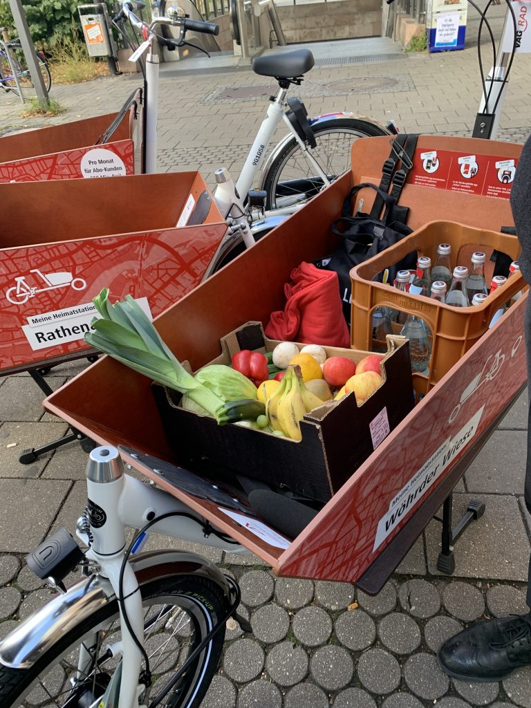 Das Foto zeigt die Transportbox eines Lastenrads. Sie ist gefüllt mit Obst, Gemüse, einem Wasserkasten, Rucksack und einer Decke.