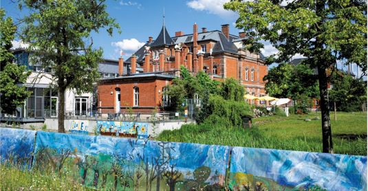 Auf dem Foto ist ein Backsteinhaus mit einem modernen Glasanbau zu sehen. Drum herum sind Bäume, Sträuche und Rasen.