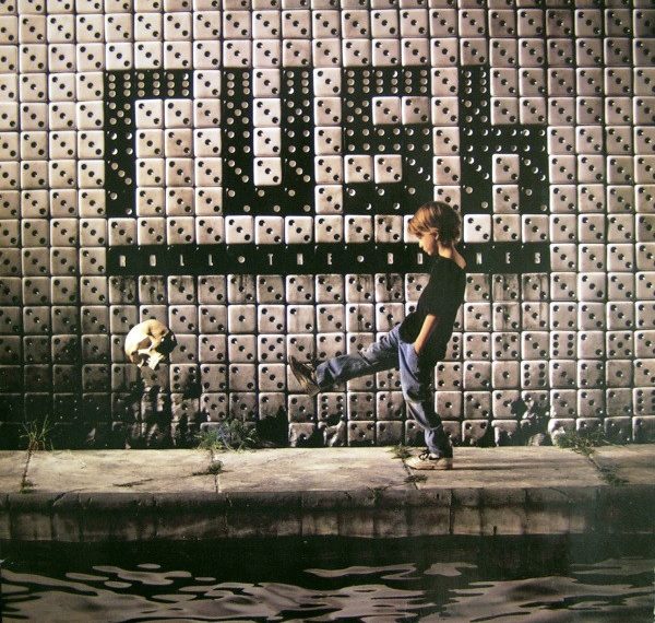 Das Albumcover "Roll The Bones" von Rush zeigt eine Wand aus weißen und schwarzen Würfeln. Die schwarzen Würfeln bilden das Wort "Rush". Vor der Wand steht ein Junge, der einen Totenkopf mit dem Fuß wegkickt.