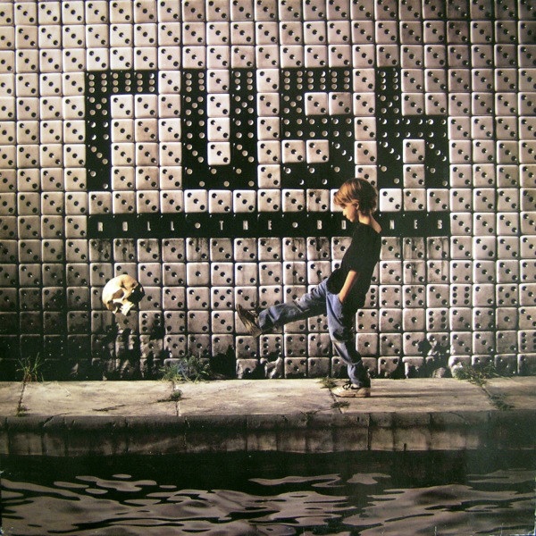 Das Albumcover "Roll The Bones" von Rush zeigt eine Wand aus weißen und schwarzen Würfeln. Die schwarzen Würfeln bilden das Wort "Rush". Vor der Wand steht ein Junge, der einen Totenkopf mit dem Fuß wegkickt.