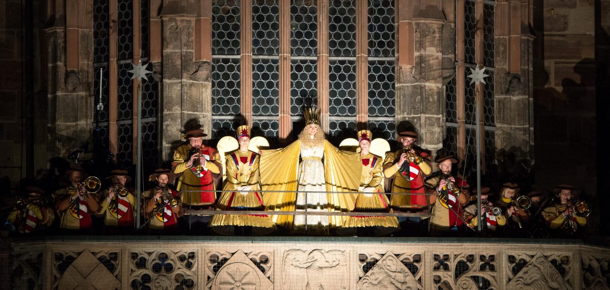 Auf dem Foto sieht man die Empore der Frauenkirche. Dort stehen mehrere Menschen. In der Mitte steht Benigna Munsi als Nürnberger Christkind und sagt den Prolog auf.