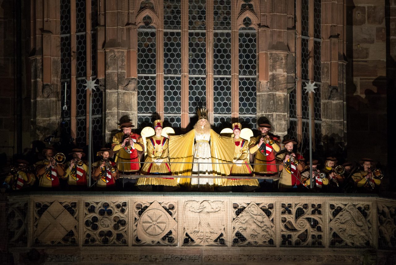 Auf dem Foto sieht man die Empore der Frauenkirche. Dort stehen mehrere Menschen. In der Mitte steht Benigna Munsi als Nürnberger Christkind und sagt den Prolog auf.