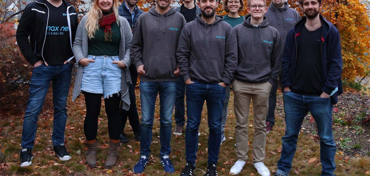 Auf dem Foto sieht man das Team von max neo vor einer gelben Hecke. Es sind acht Männer, zwei Frauen und eine Hündin zu sehen.