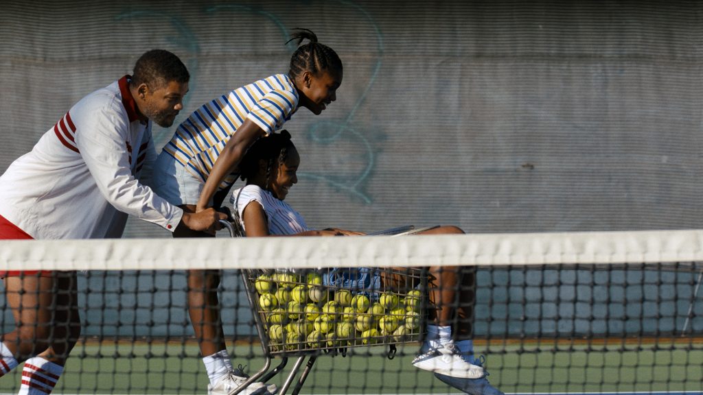 Will Smith als Richard Williams schiebt einen Einkaufswagen voller Tennisbälle über ein Tennisfeld. Auf dem Wagen sitzen die zwei Schauspielerinnen, die Venus und Serena Williams verkörpern.