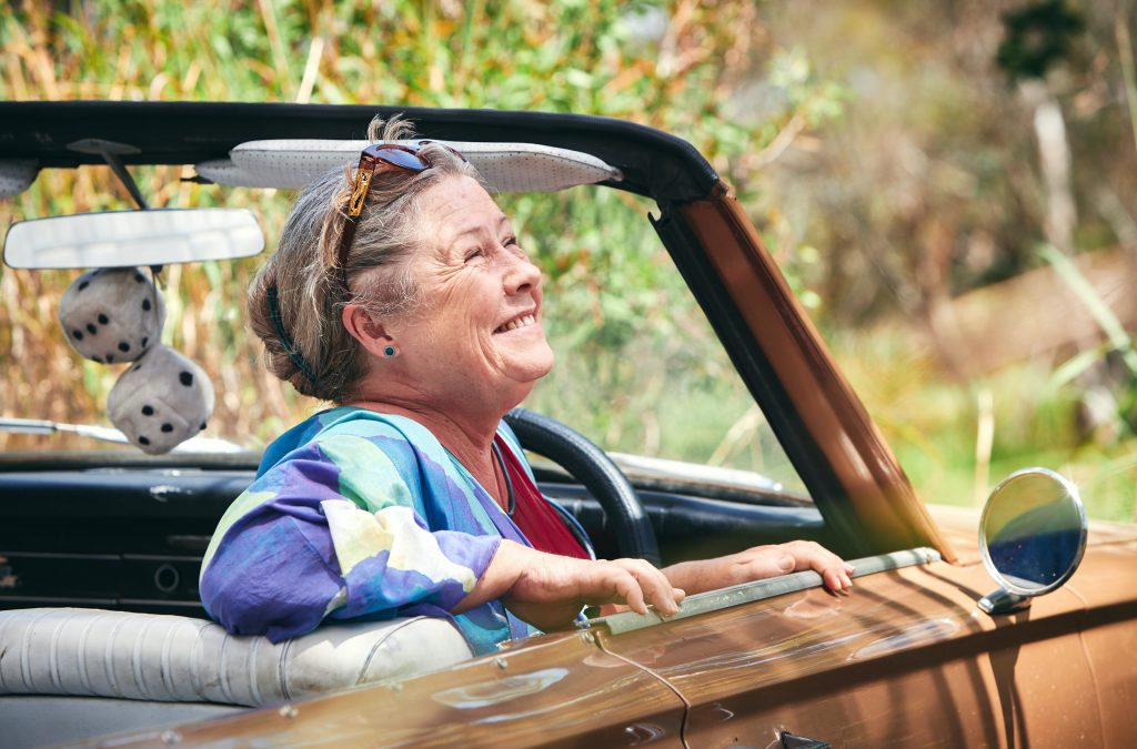 Noni Huzlehurst sitzt als June in einem Oldtimer und lacht Richtung Himmel.