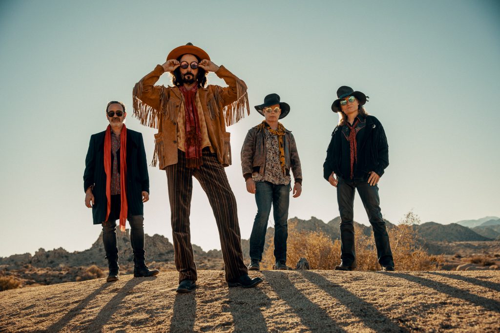 Das Foto zeigt die Band Mike Campbell & The Dirty Knobs. Die vier Männer stehen in einer Wüste und haben Sonnenbrillen auf. Im Hintergrund sind Felsformationen zu sehen.