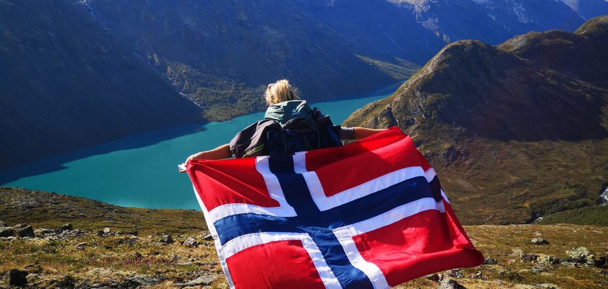 Celine Pickel steht mit dem Rücken zur Kamera und hält eine norwegische Flagge in den Händen. Sie steht auf einem Berg, im Hintergrund sind ein Fjord und Berge zu sehen.