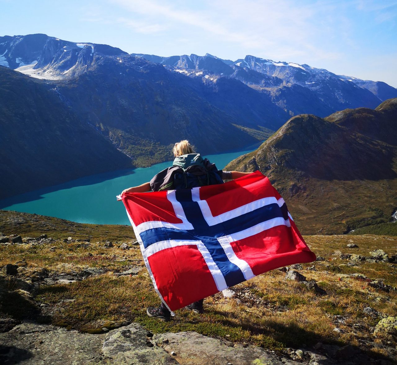 Celine Pickel steht mit dem Rücken zur Kamera und hält eine norwegische Flagge in den Händen. Sie steht auf einem Berg, im Hintergrund sind ein Fjord und Berge zu sehen.