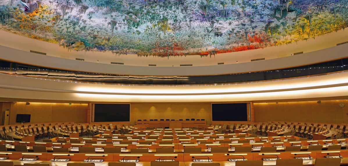 Das Foto zeigt den Saal des Menschenrechtsrats in Genf. Man sieht ganz viele Tische mit Stühlen und Mikrofonen. An der Decke ist ein buntes 3D-Gemälde zu sehen.