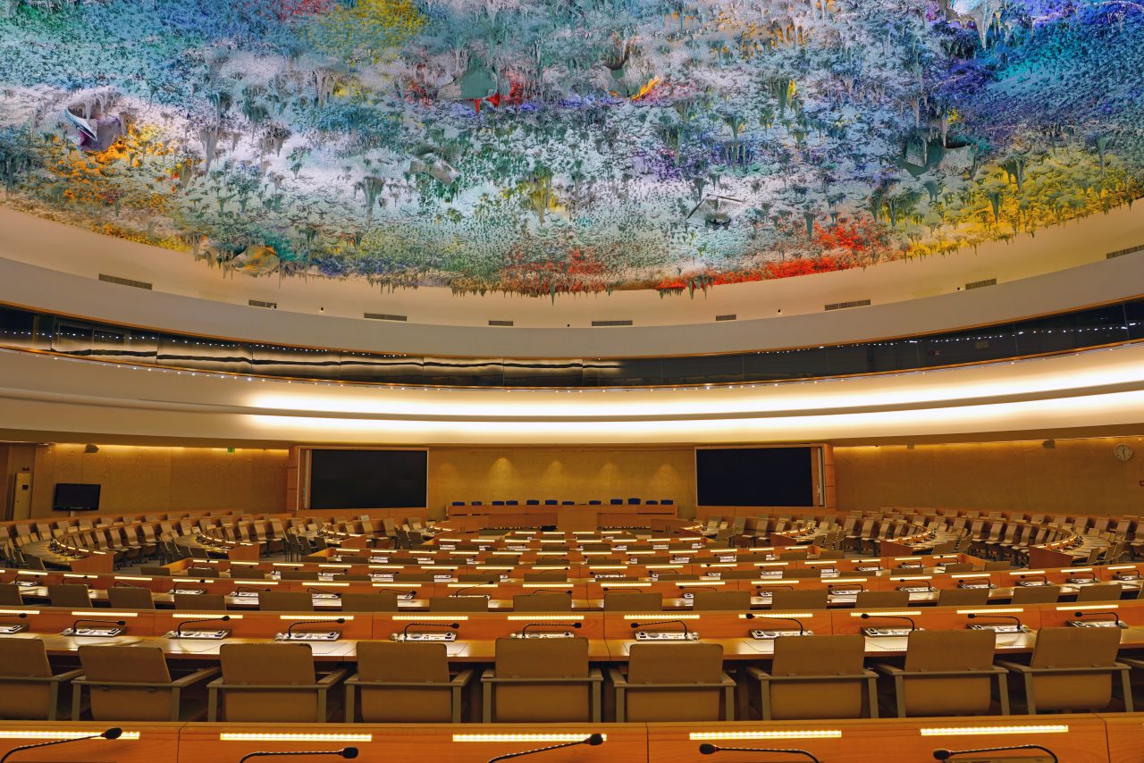 Das Foto zeigt den Saal des Menschenrechtsrats in Genf. Man sieht ganz viele Tische mit Stühlen und Mikrofonen. An der Decke ist ein buntes 3D-Gemälde zu sehen.