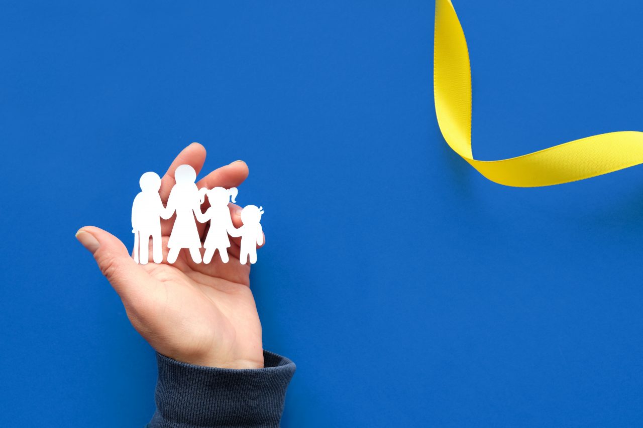 Der Hintergrund des Bildes ist blau. Rechts oben ist ein gelbes Band zu sehen. In der Mitte befindet sich eine Hand, die vier weiße Figuren in der Hand hält, die eine Familie darstellen sollen.