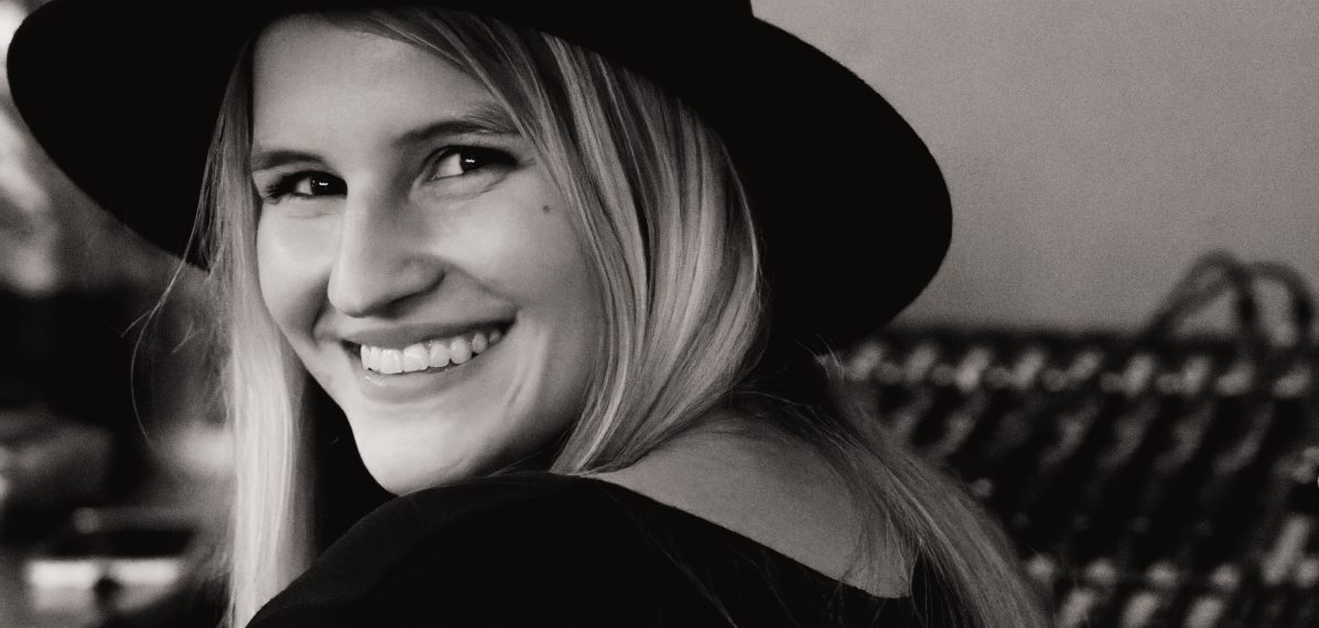 Das Schwarz-Weiß-Foto zeigt die Comedienne Julia Brandner, die einen Hut auf dem Kopf hat und in die Kamera lächelt.