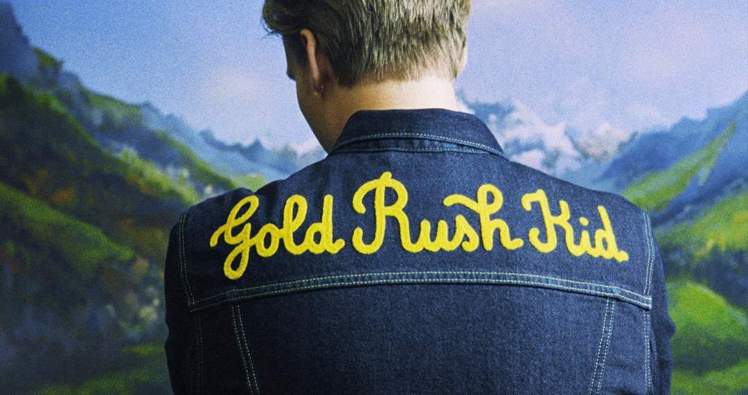 Das Albumcover "Gold Rush Kid" von George Ezra zeigt den Musiker von hinten. Er trägt eine Jeansjacke mit dem aufgestickten Albumtitel. Im Hintergrund sind ein See und Berge zu sehen.