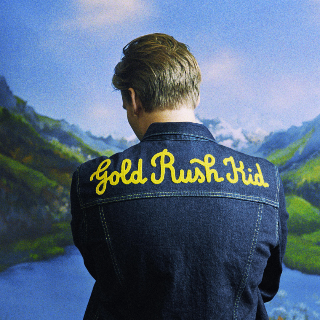 Das Albumcover "Gold Rush Kid" von George Ezra zeigt den Musiker von hinten. Er trägt eine Jeansjacke mit dem aufgestickten Albumtitel. Im Hintergrund sind ein See und Berge zu sehen.