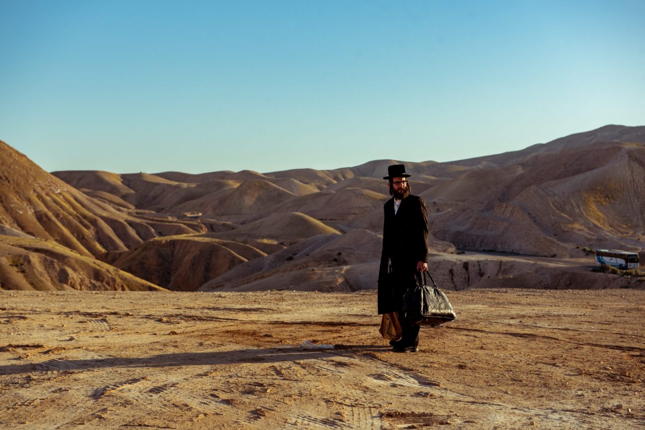 Das Foto ist eine Momentaufnahme aus dem Film "Nicht ganz koscher". Es zeigt einen Mann mit Taschen in der Hand, der in einer wüstenähnlichen Hügellandschaft steht.