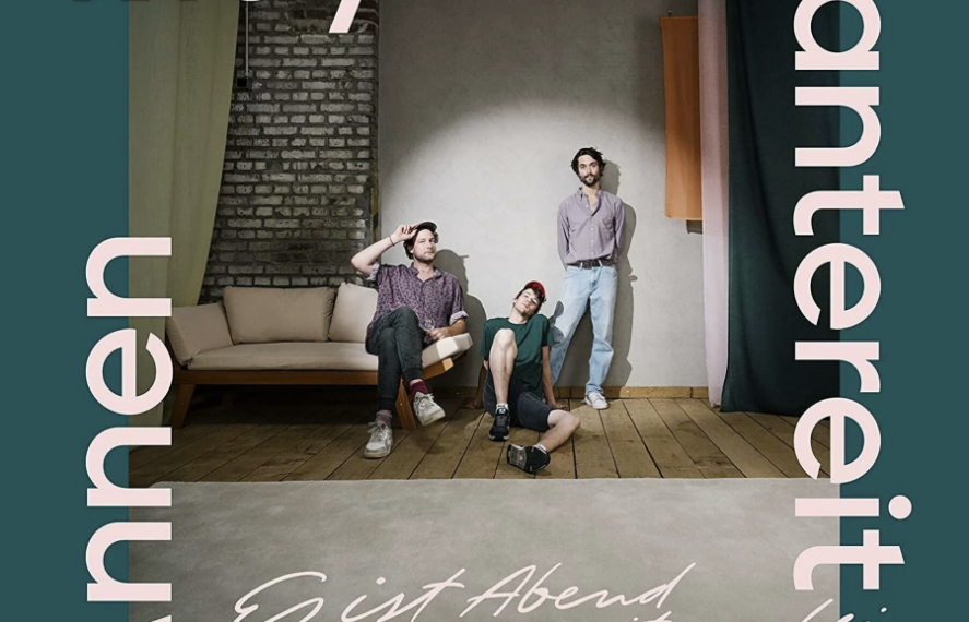 Das Albumcover "Es ist Abend und wir sitzen bei mir" von AnnenMayKantereit zeigt das Trio, wie sie in ihrem Proberaum sitzen oder stehen und in die Kamera schauen. Umrahmt ist das Foto von einem dunkelgrünen Rahmen.