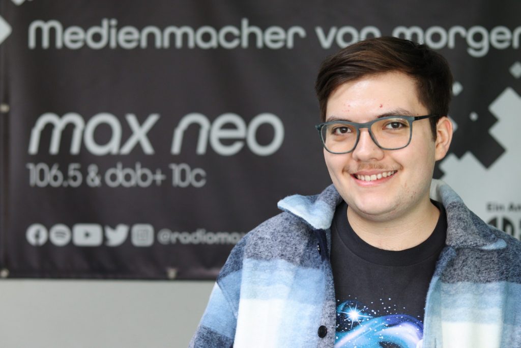 Das Foto zeigt den Praktikanten Leonardo Garcia im Porträt vor dem max neo Banner.