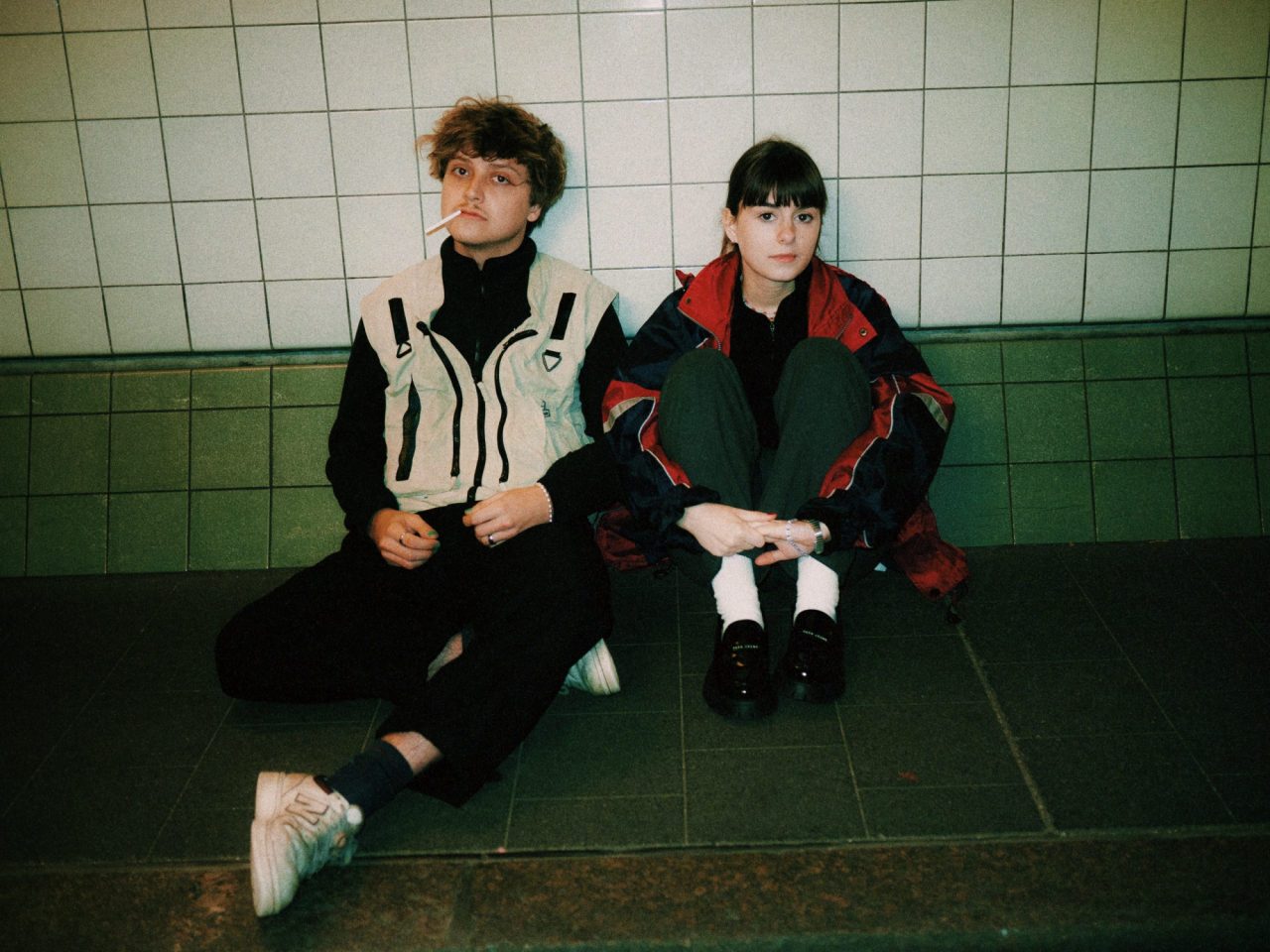 Das Foto zeigt das Indiepopduo Lena & Linus. Die beiden sitzen auf dem Boden. Um sie herum sind der Boden und die Wand gefliest.