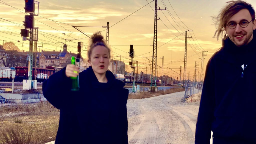 Das Foto zeigt die zwei Bandmitglieder von "Nun flog Dr. Bert Rabe", die an einem Bahnhof im Sonnenuntergang stehen. Mila hält ein Bier in die Luft.