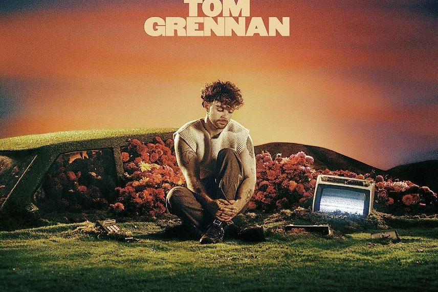 Das Albumcover "What Ifs & Maybes" von Tom Grennan zeigt den Musiker, wie er mit nachdenklichem Blick auf einer Wiese sitzt. Der Himmel im Hintergrund ist blau, violett, rosa, orange und gelb.