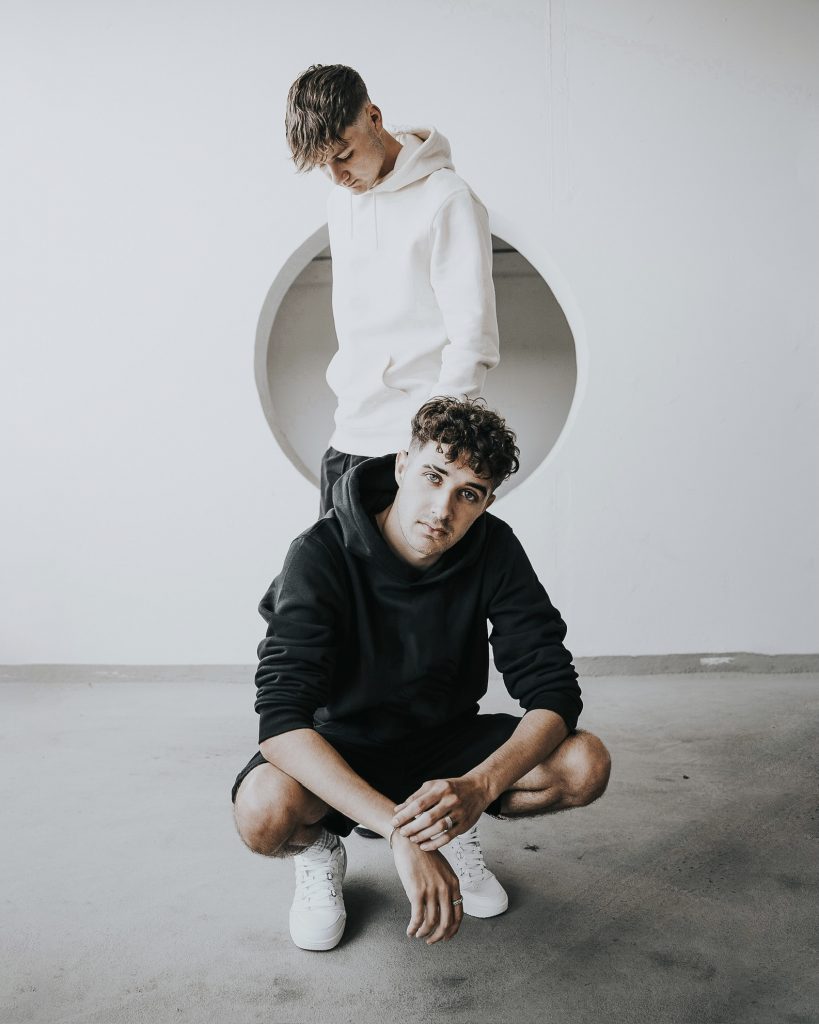 Das Foto zeigt die beiden DJs Luca-Dante Spadafora und Niklas Dee. Niklas steht vor einer weißen Wand, vor ihm ist Luca in der Hocke.