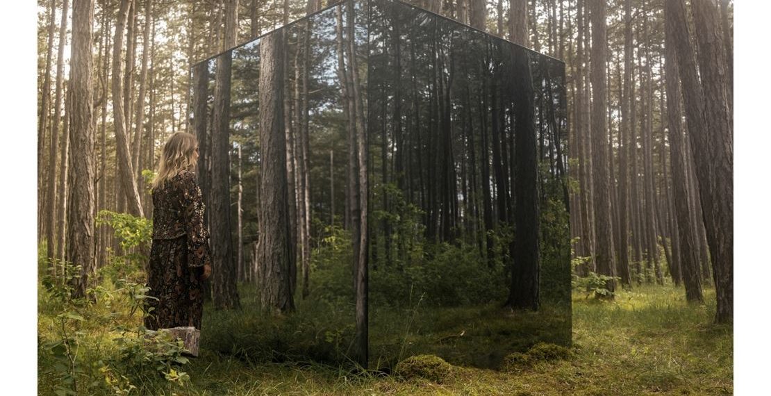 Das Albumcover "I Feel Alone These Days" von Avec zeigt die Musikerin in einem Wald. Sie steht vor einem großen, verspiegelten Würfel.