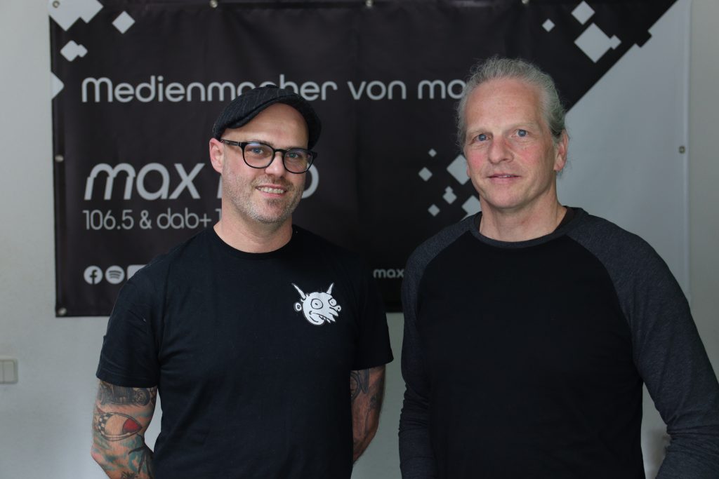 Das Foto zeigt zwei Mitglieder der Band ØL. Sie stehen vor dem max neo Banner.