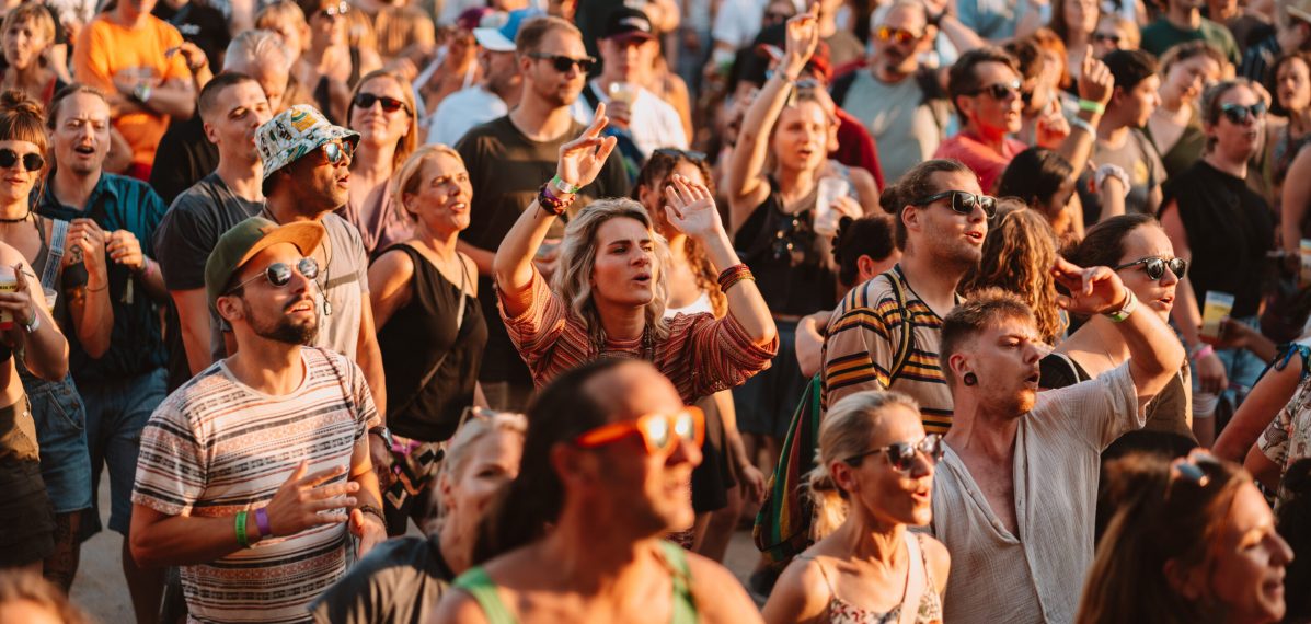 Das Foto zeigt eine Gruppe von Menschen, die beim AKF Festival 2024 tanzen. Sie haben sommerliche Klamotten an, das Bild ist in einem warmen Setting, vermutlich bei Sonnenuntergang aufgenommen