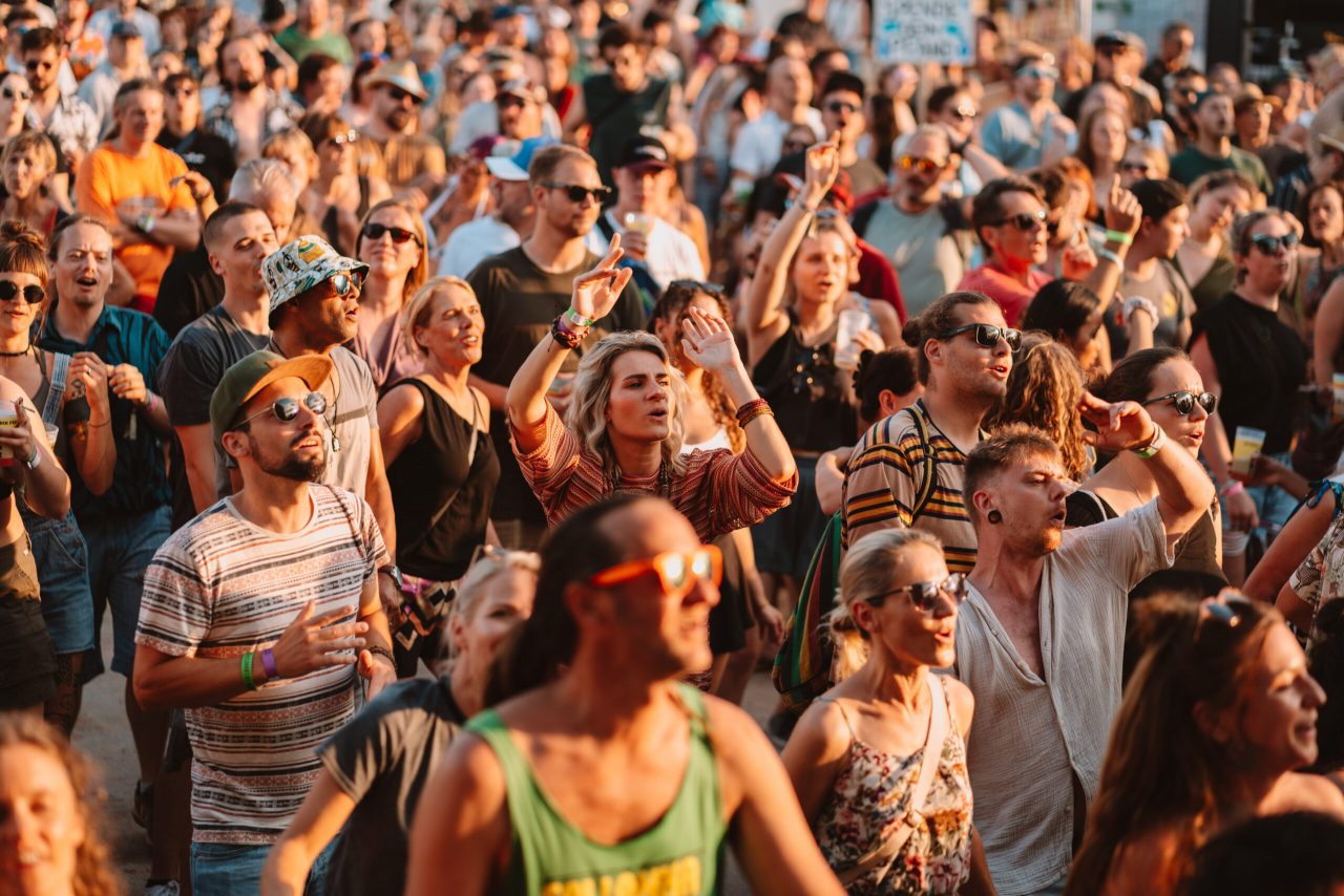 Das Foto zeigt eine Gruppe von Menschen, die beim AKF Festival 2024 tanzen. Sie haben sommerliche Klamotten an, das Bild ist in einem warmen Setting, vermutlich bei Sonnenuntergang aufgenommen