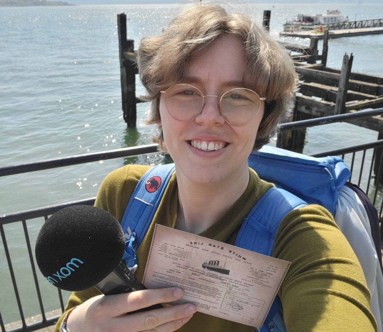 Das Foto zeigt Lena Schnelle mit Mikrofon und Fahrkarte in der Hand vor dem Meer.