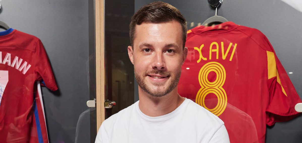 Der Content Creator Lukas Hösch, der unter dem Namen Badeschlappen bekannt ist in einer Spielerumkleide eines Fußballvereins. Im Hintergrund ist das rote Trikot des Fußballers Xavi und die Nummer 8 zu sehen. Es handelt sich dabei um das spanische Nationaltrikot. Lukas trägt ein weißes, kurzärmeliges Shirt und eine dunkle Hose.