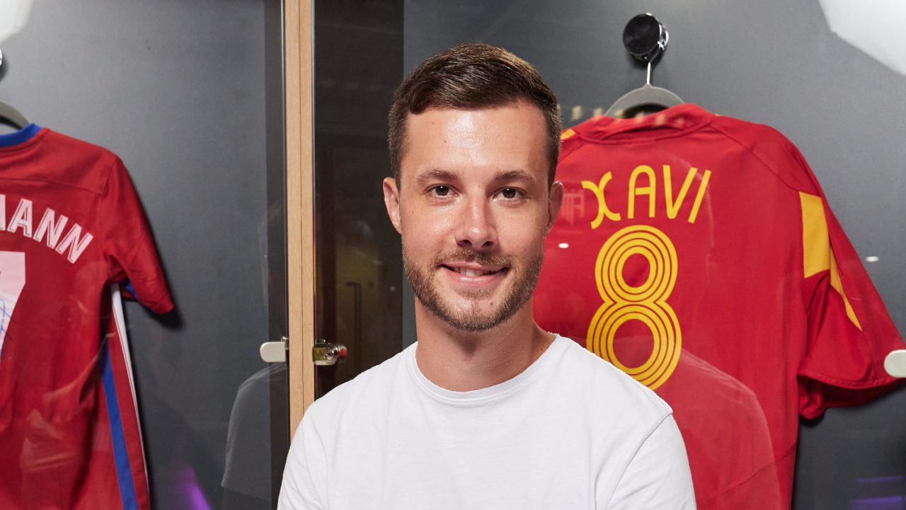 Der Content Creator Lukas Hösch, der unter dem Namen Badeschlappen bekannt ist in einer Spielerumkleide eines Fußballvereins. Im Hintergrund ist das rote Trikot des Fußballers Xavi und die Nummer 8 zu sehen. Es handelt sich dabei um das spanische Nationaltrikot. Lukas trägt ein weißes, kurzärmeliges Shirt und eine dunkle Hose.