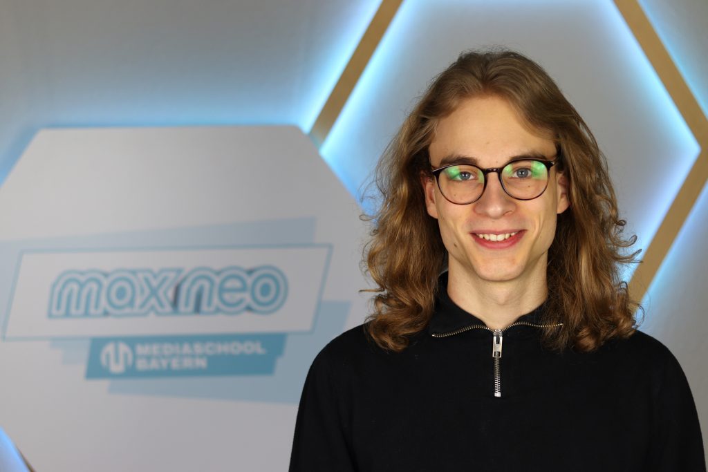 Das Foto zeigt den Praktikanten Robin Pfeiffer im Porträt vor dem max neo Banner.