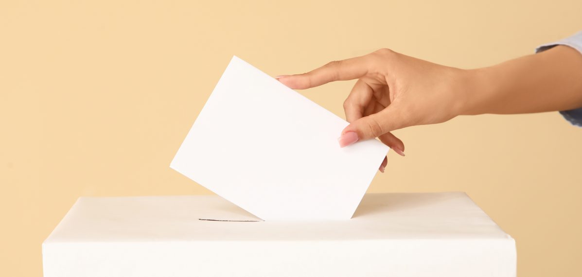 Das Foto zeigt eine Hand, die einen Wahlzettel in eine Urne schmeißt.